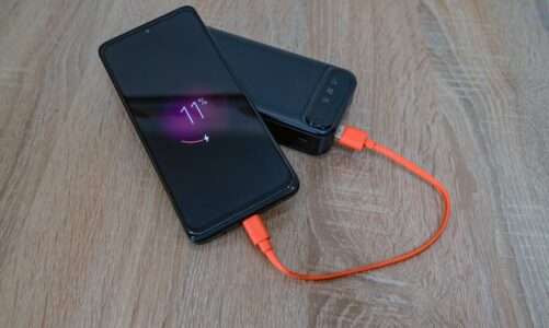 Czarny smartfon ładowany przy użyciu pomarańczowego kabla z powerbanku na drewnianym stole.
