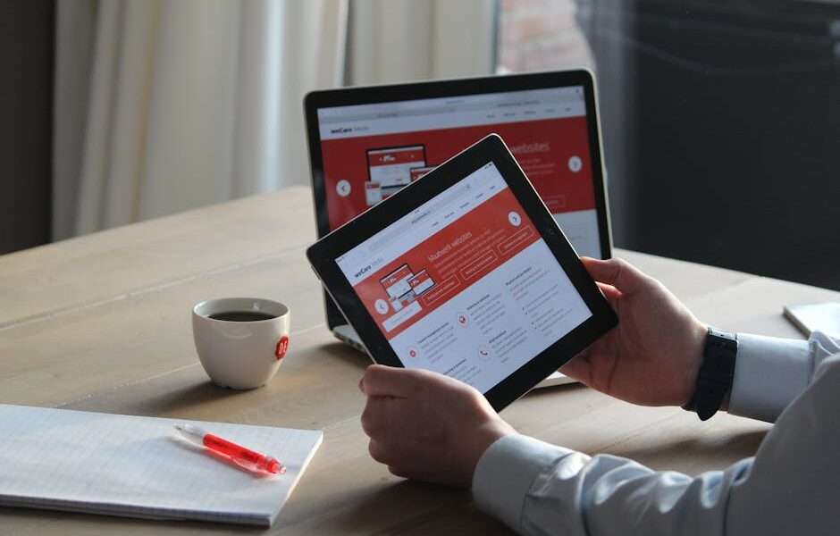 Osoba trzymająca tablet z otwartą stroną internetową obok laptopa i notatnika.