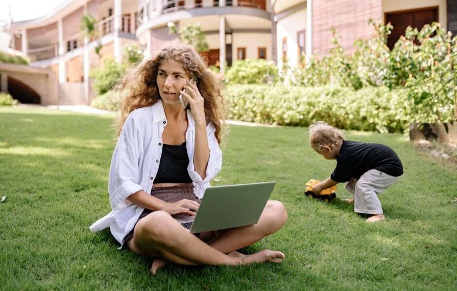 kobieta rozmawiająca przez telefon i trzymająca laptopa na kolanach w trakcie opieki nad dzieckiem w ogrodzie